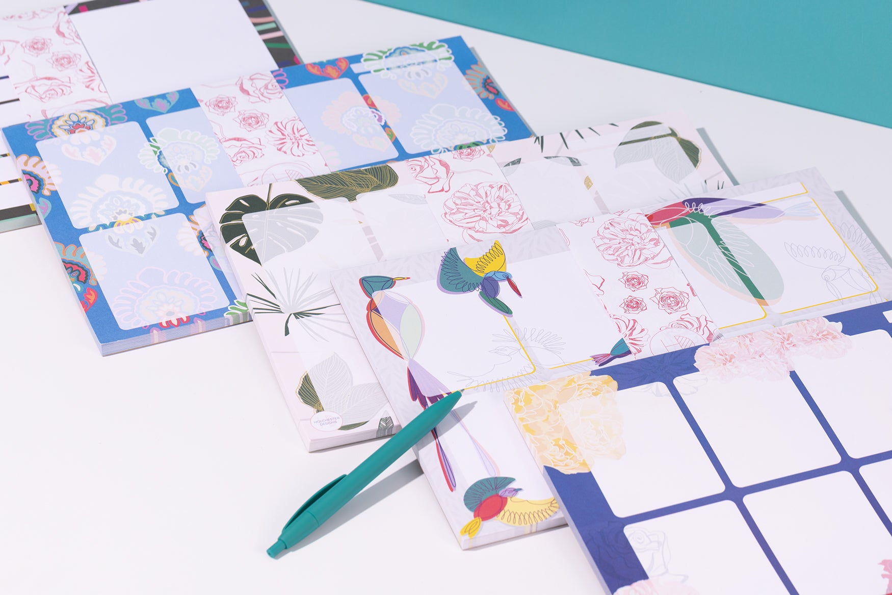 Stack of 5 different Planner pads, leaning on each other from bottom right to top left.  They are all wrapped with belly bands around them and are on a white desk with a teal pen alongside.