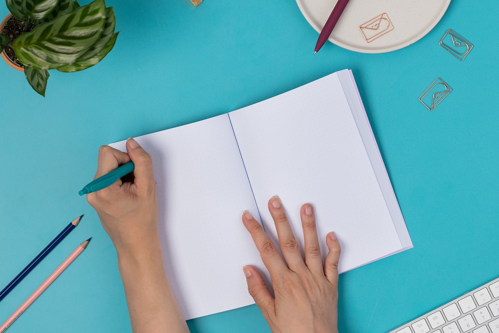 A5 Dot Grid notebook being held open. A hand on the left holding a teal pen is writing on the topleft page, with a right hand holding the book in the middle. it is leaning on a teal desk with a white keyboard and paperclips around it.