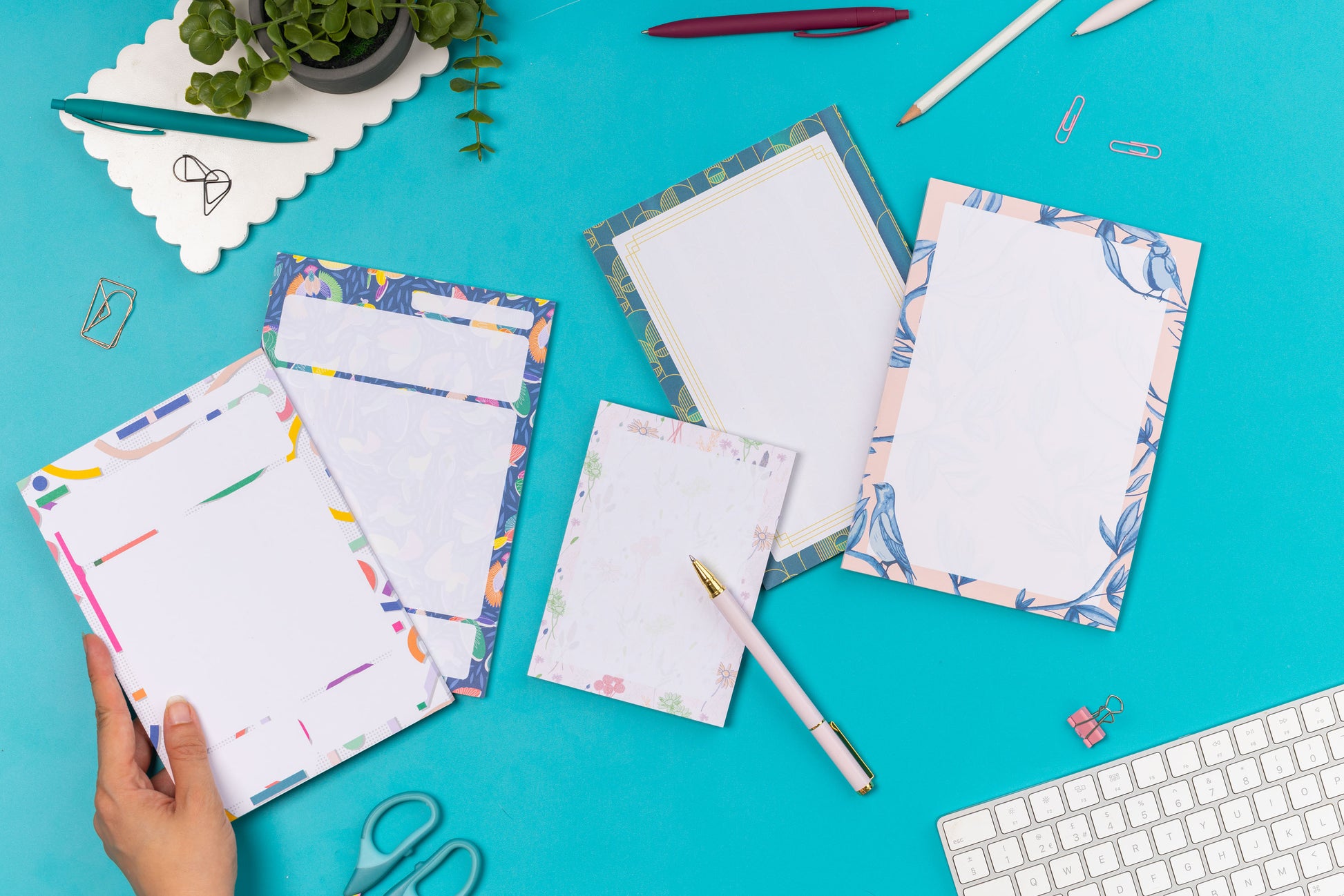 4 A5 notepads (Cutouts, Paradise, Deco Delights and Brighton Birds) are flat in a row with the A6 Home Grown pad, on a teal desk with pens, paperclips and a white keyboard around them.  There is a hand holding the cutouts pad in the bottom left of the image.