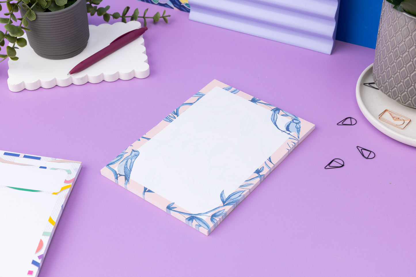 Brighton Birds A5 Notepad - Blush pink background with shades of blue bird and plant hand-painted motifs - is on a lilac desk, next to a Cutouts notepad (front left out of focus) and scattered paperclips.