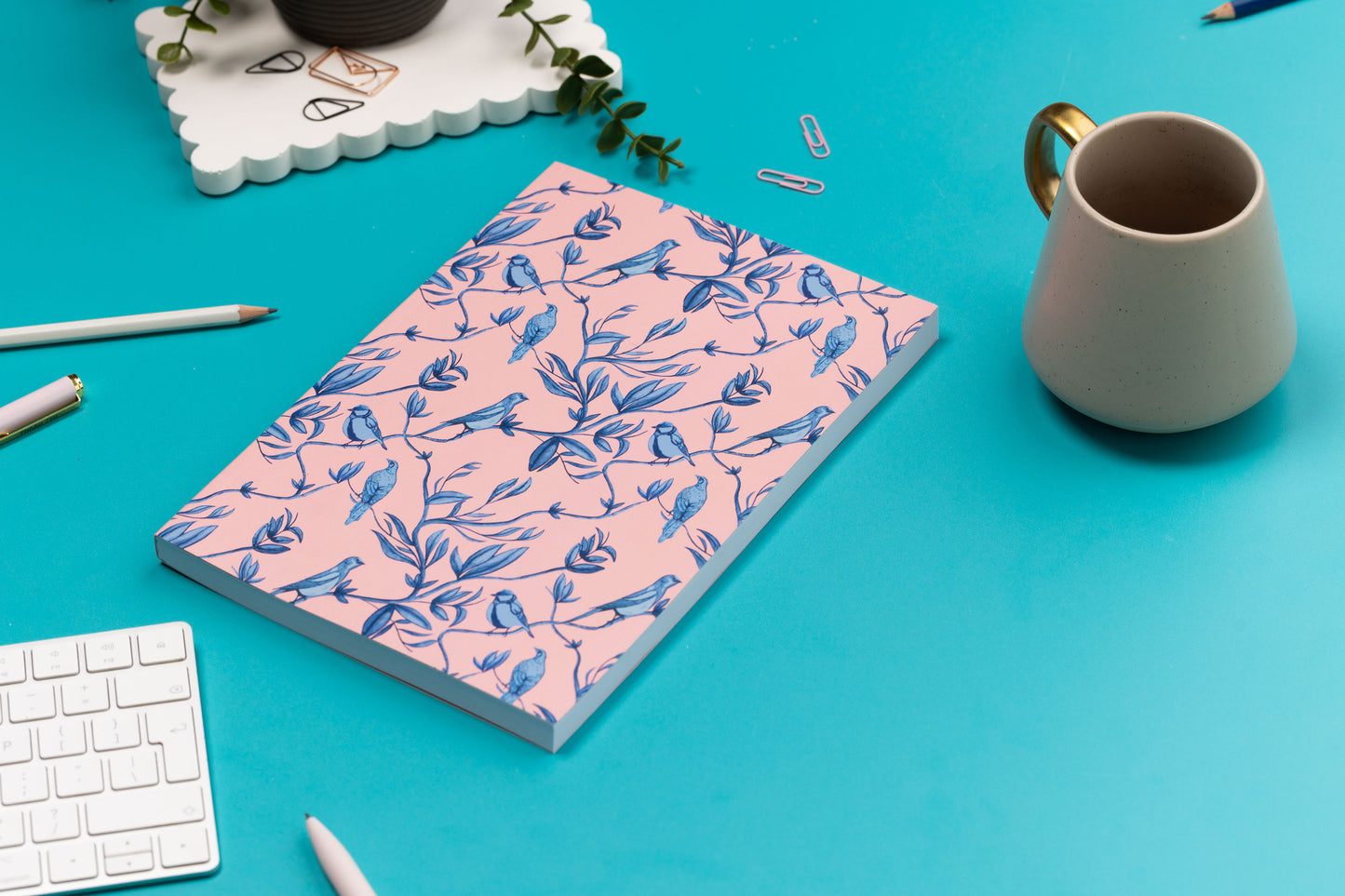 Brighton Birds B5 Dot Grid Notebook is central, on a teal desk with a white keyboard, grey mug, scattered pens and a small plant around it.  