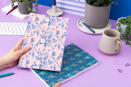 The Pair of B5 dot grid notebooks - Deco Delights below and Brighton Birds being held by a hand to its left.  They are on a lilac desk with a white keyboard, two small plant, a mug and scattered pens around them.