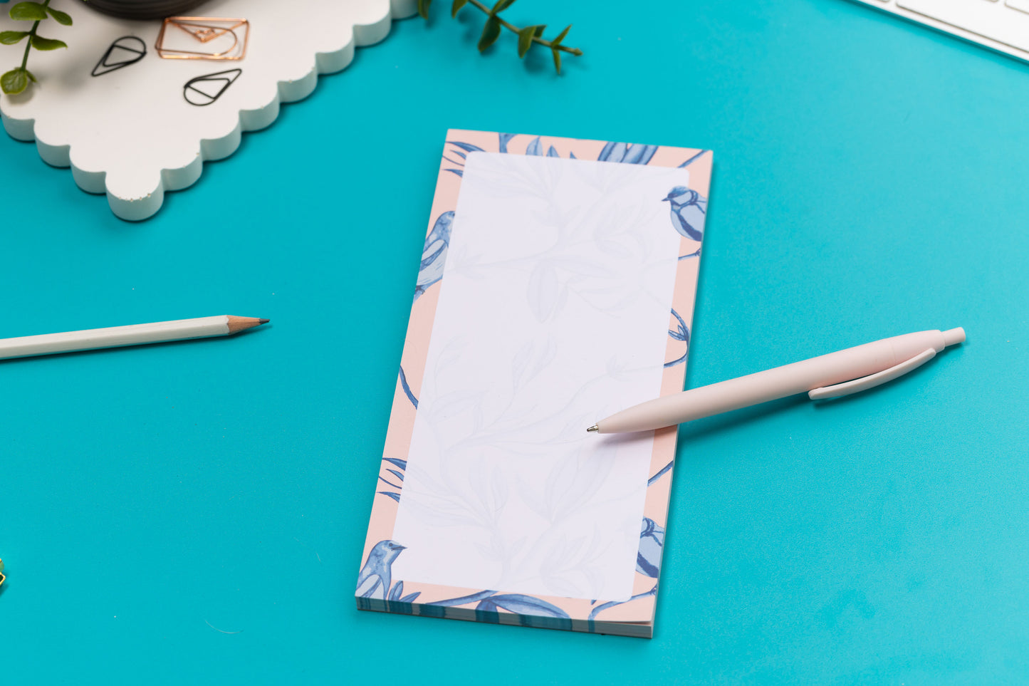 Brighton Birds List pad - Blush pink background with shades of blue bird and plant hand-painted motifs - is on a teal desk, next to a white pen, white keyboard and scattered paperclips.