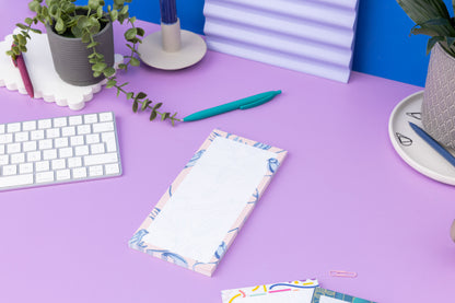 Brighton Birds List pad - Blush pink background with shades of blue bird and plant hand-painted motifs - is on a lilac desk, next to a white pen, white keyboard and scattered paperclips.