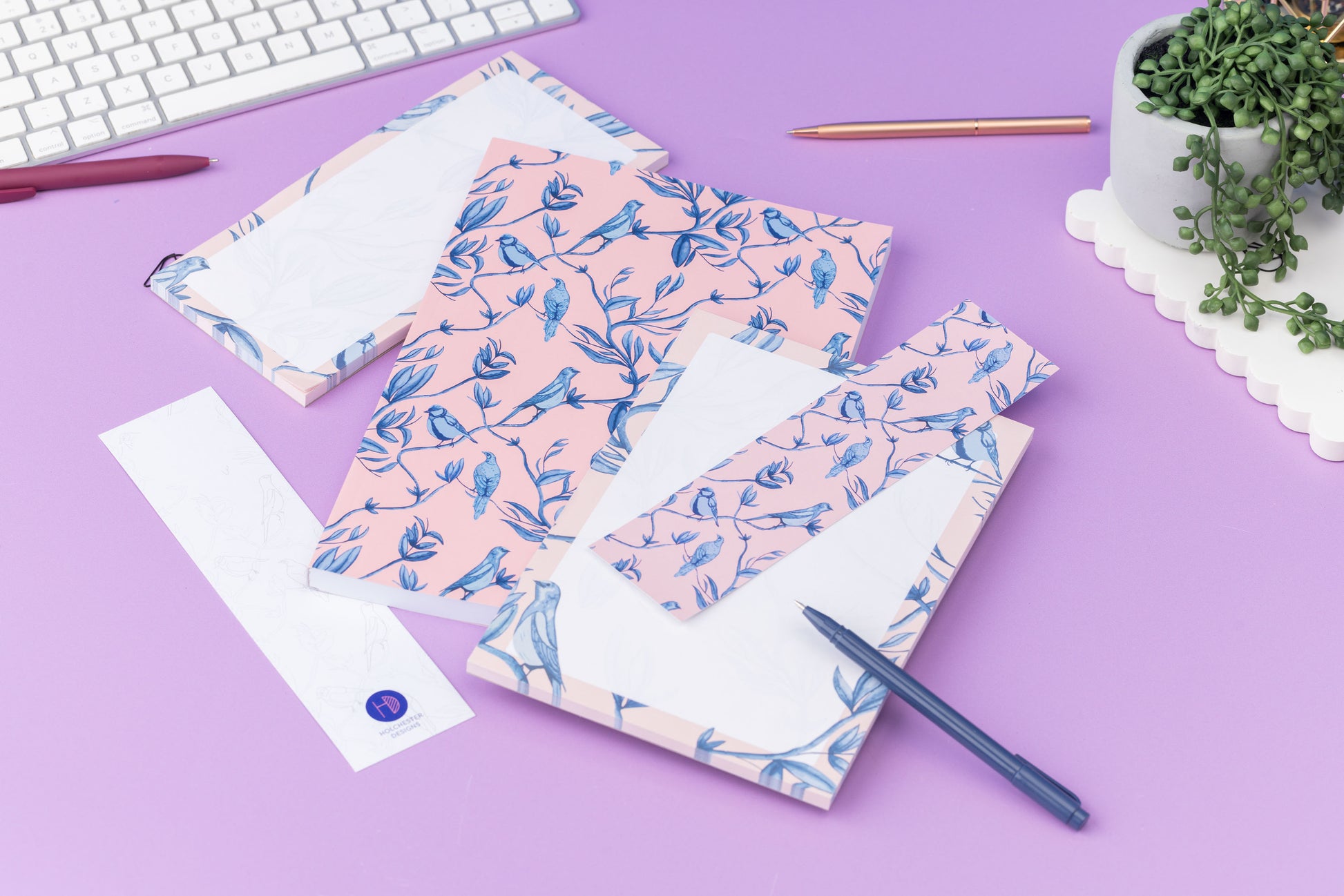 Brighton Birds Full Colleciton - B5 Notebook, List pad, A5 Notepad and 2 double-sided bookmarks (one side up each) - is on a lilac desk, with a white keyboard, pens and a small pot plant around them.