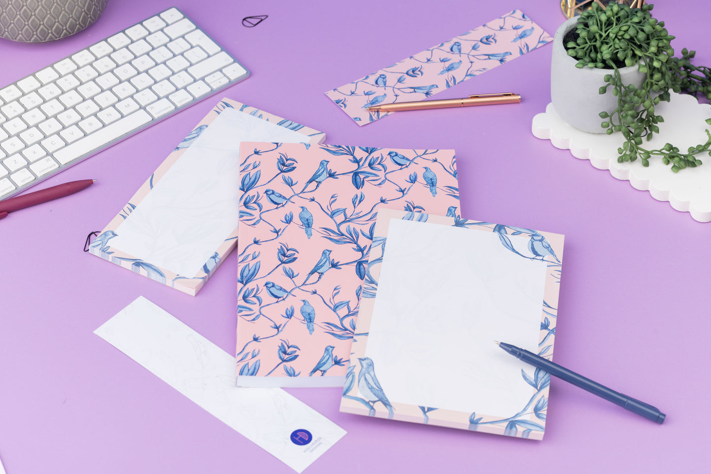 Brighton Birds Full Colleciton - B5 Notebook, List pad, A5 Notepad and 2 double-sided bookmarks (one side up each) - is on a lilac desk, with a white keyboard, pens and a small pot plant around them.