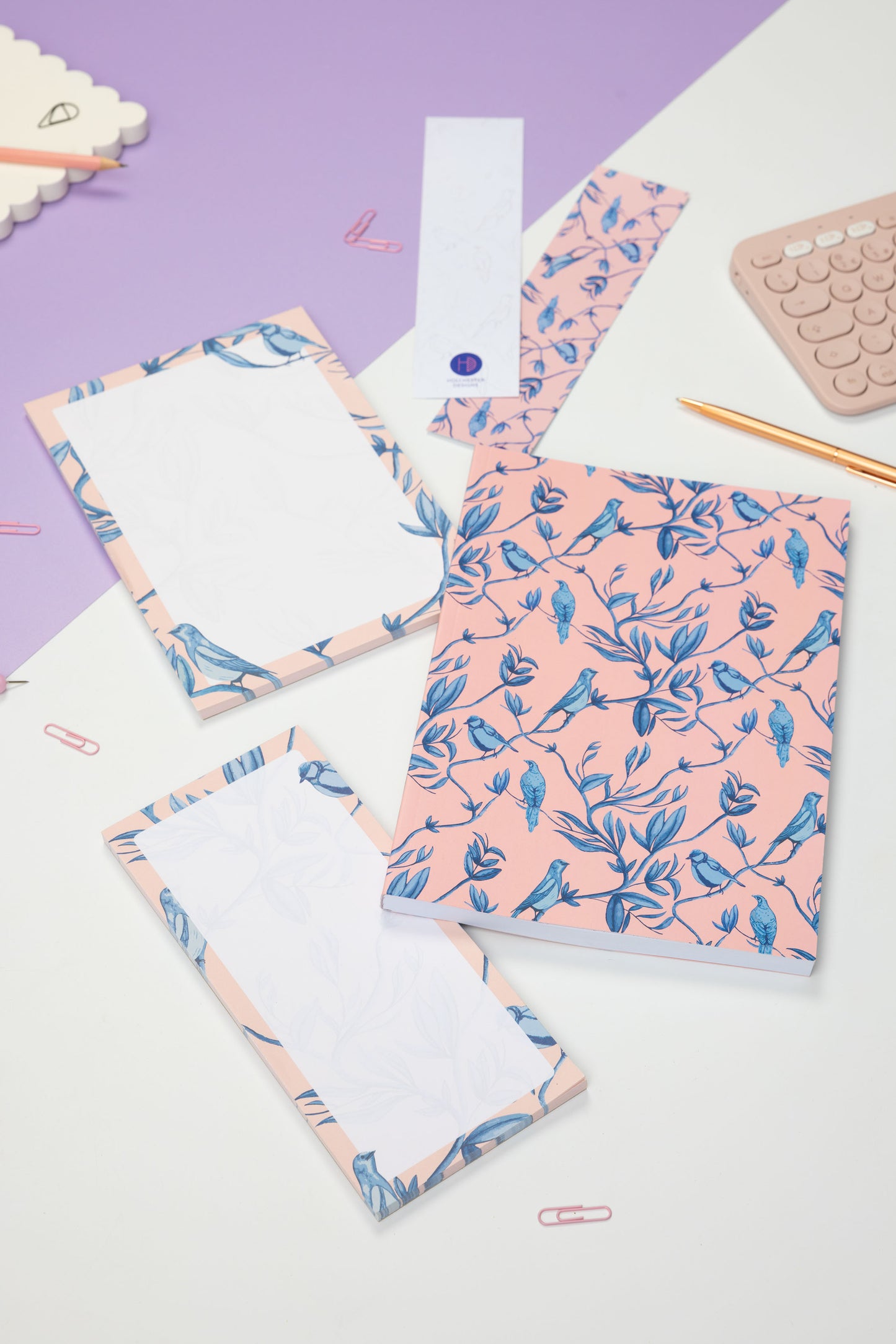 Brighton Birds Full Colleciton - B5 Notebook, List pad, A5 Notepad and 2 double-sided bookmarks (one side up each) - is on a white and lilac desk, with a blush pink keyboard and pink paperclips scattered around them.