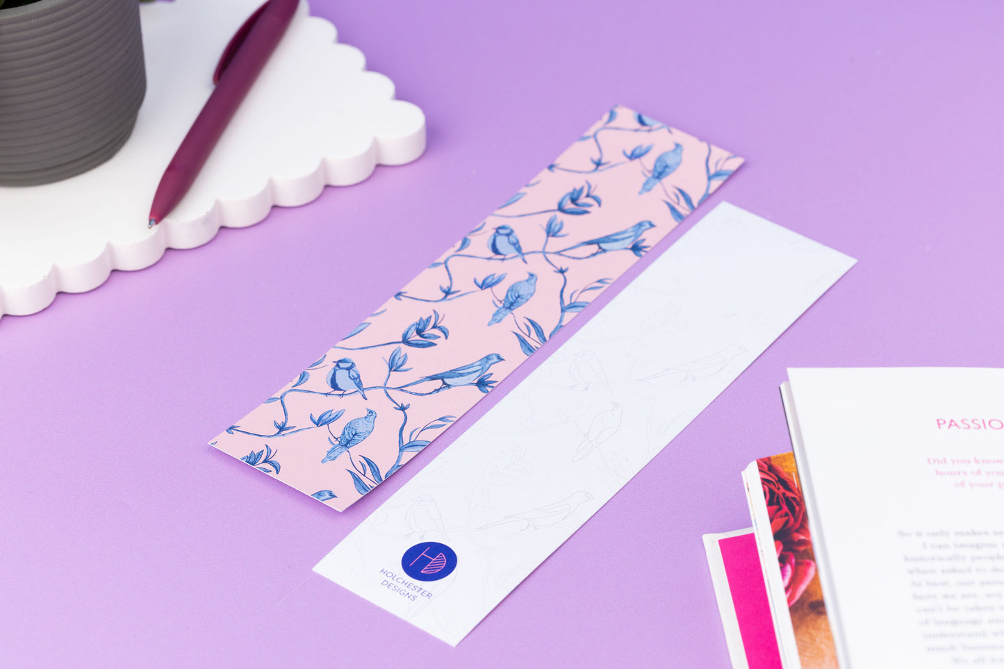 Two of the Brighton Birds double-sided bookmarks, on a lilac desk with a book open to the bottom right corner.  One side features the full-colour patterned side of the Brighton Birds pattern with the blush pink background and shades of blue branch, leaf and bird hand-painted motifs.  The other side is the light-grey line drawing.  