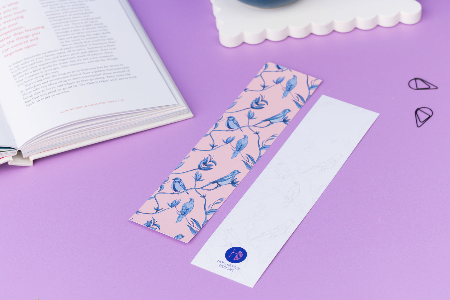 Two of the Brighton Birds double-sided bookmarks, on a lilac desk with a book open to the top left corner.  One side features the full-colour patterned side of the Brighton Birds pattern with the blush pink background and shades of blue branch, leaf and bird hand-painted motifs.  The other side is the light-grey line drawing.  