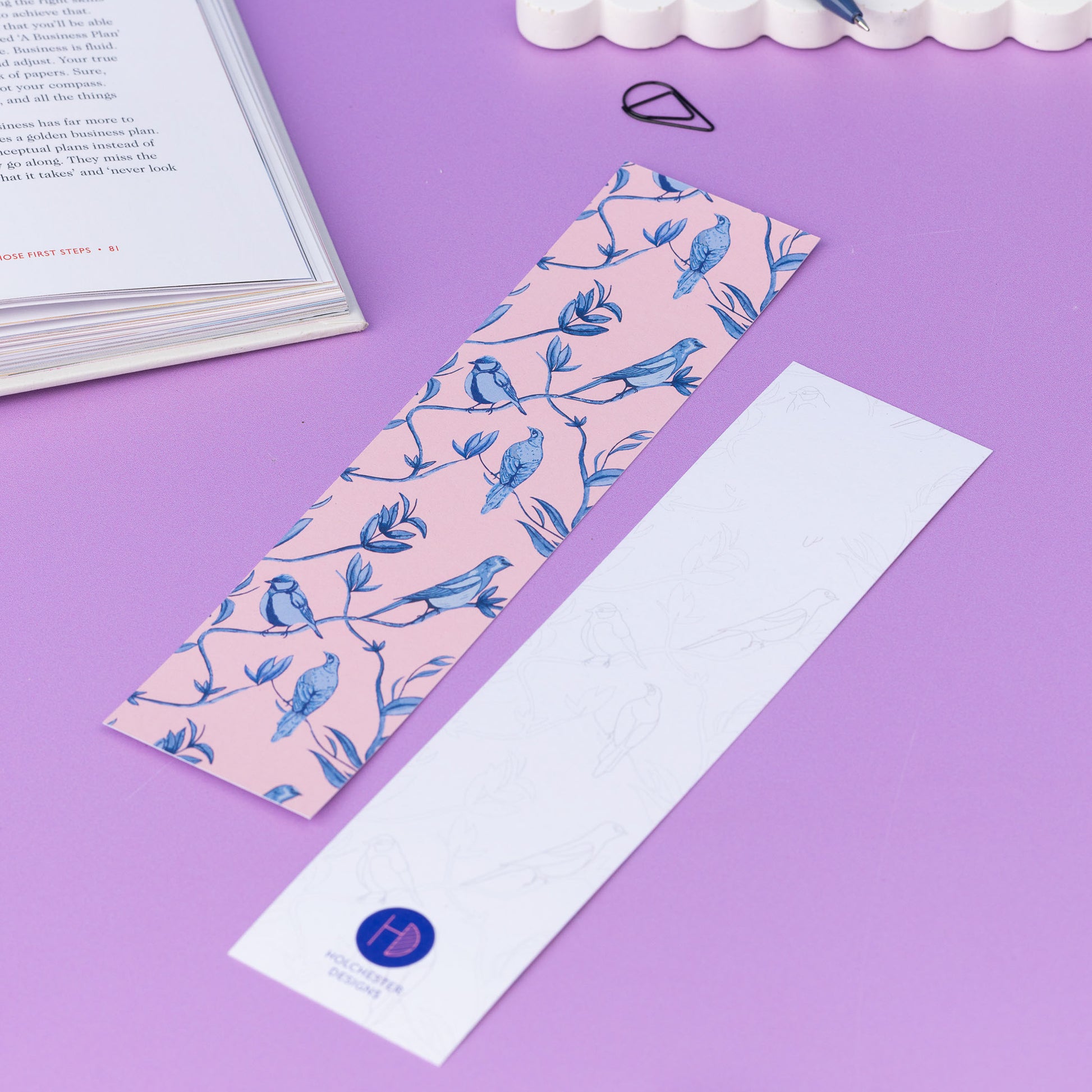 Two of the Brighton Birds double-sided bookmarks, on a lilac desk with a book open to the top left corner.  One side features the full-colour patterned side of the Brighton Birds pattern with the blush pink background and shades of blue branch, leaf and bird hand-painted motifs.  The other side is the light-grey line drawing.