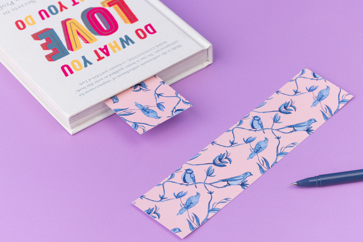 Brighton Birds double-sided bookmark is full-colour pattern side up on a lilac desk.  To its left is a closed book which has the same bookmark sticking out the top.