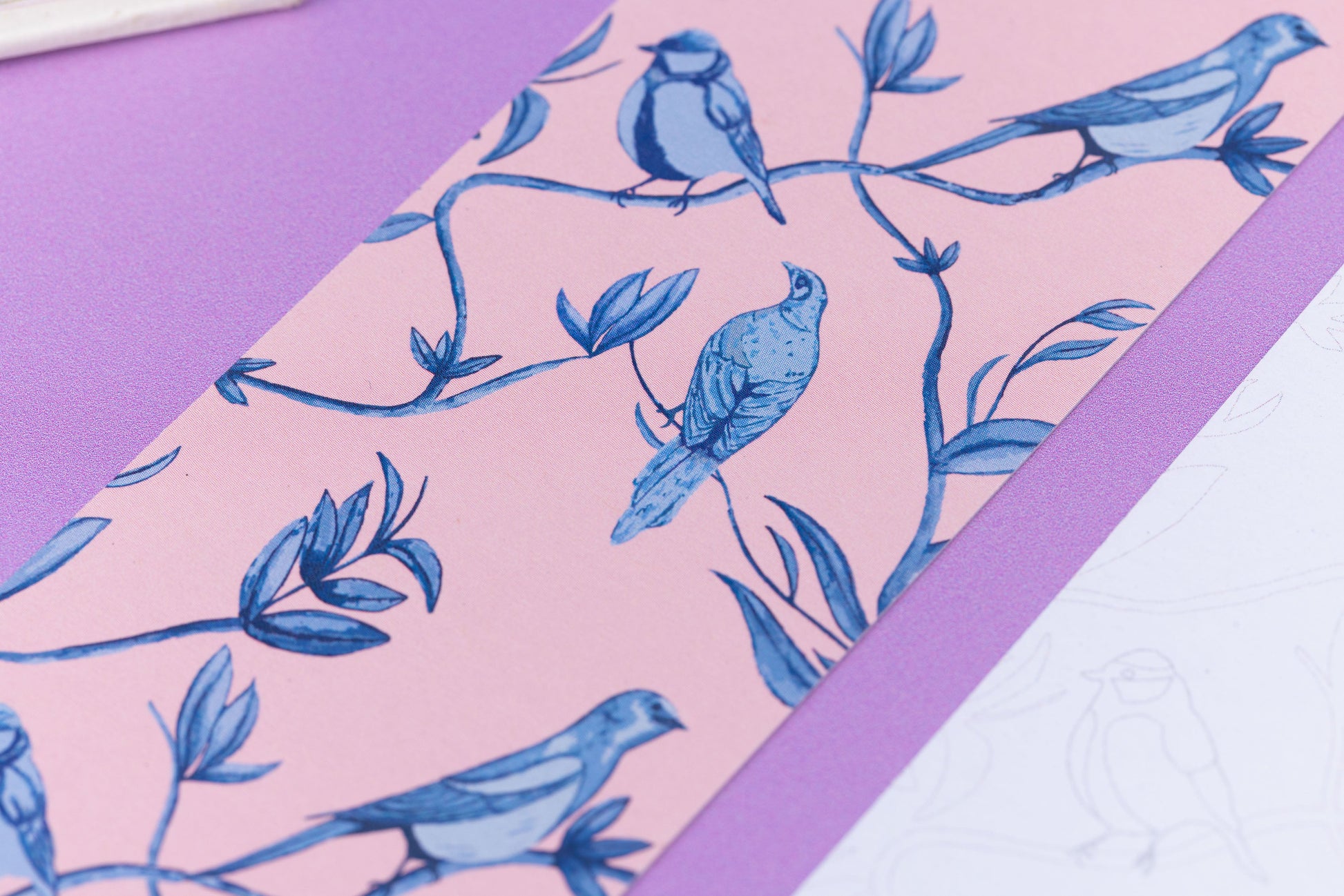 Close up of the full-colour patterned side of the Brighton Birds bookmark with a blush pink background and the blue leaves, branches and birds illustrations.  Next to is is an out of focus corner of the bookmark's other side. They are on a lilac desk with an open book in the top left corner.