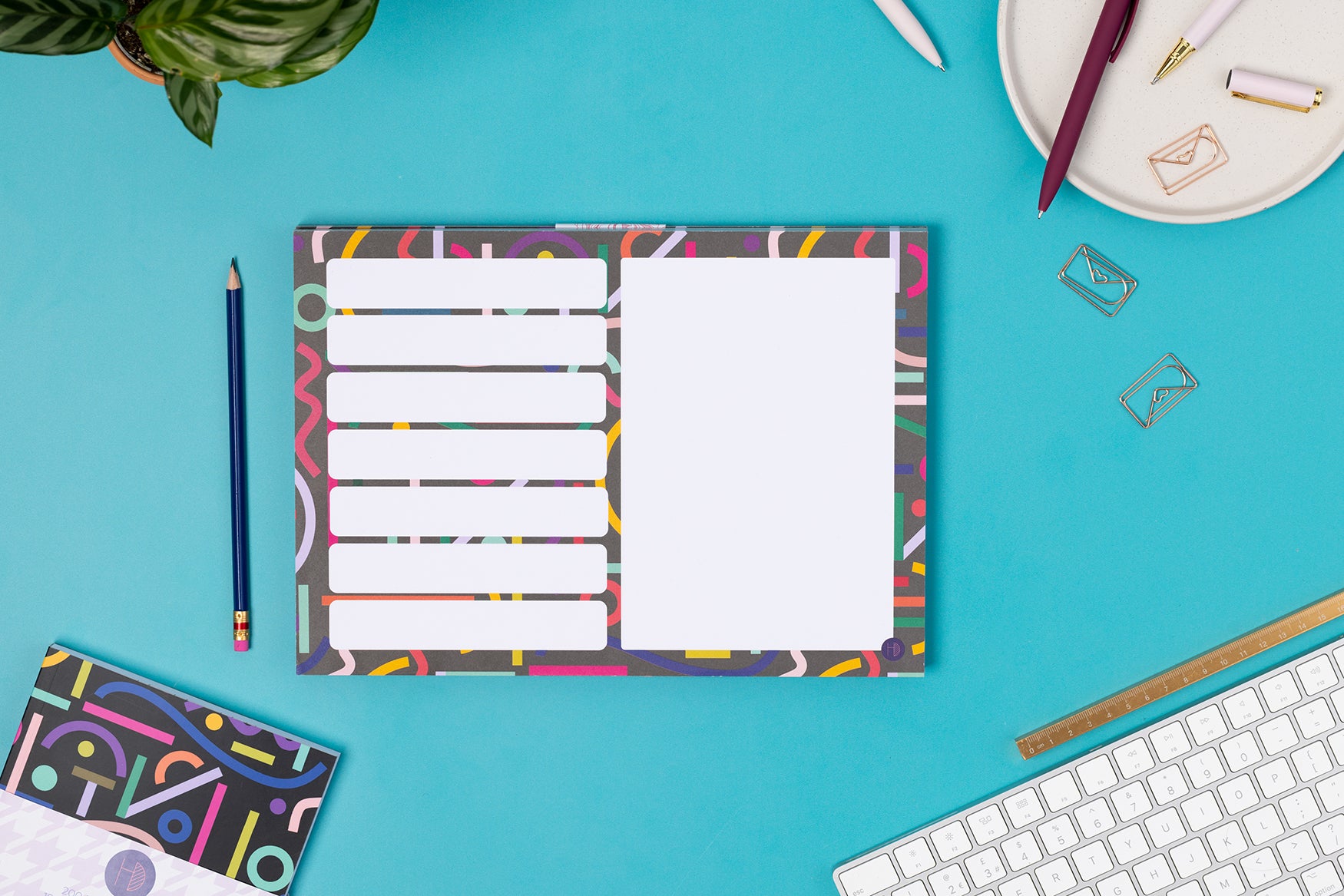 Cutouts A4 Meal/Shopping List Weekly planner pad is on a teal desk with a white keyboard and paperclips around it. There is a Cutouts A5 Notebook to the bottom right corner.