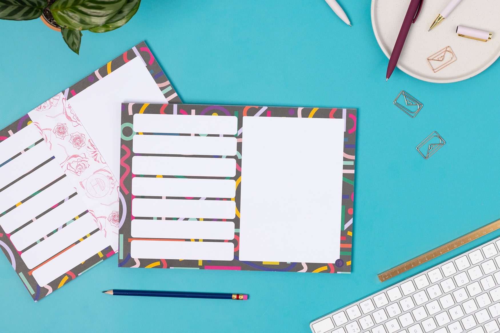 Cutouts A4 Meal/Shopping List Weekly planner pad is leaning on another pad with a belly band.  They are on a teal desk with a white keyboard and paperclips around it.