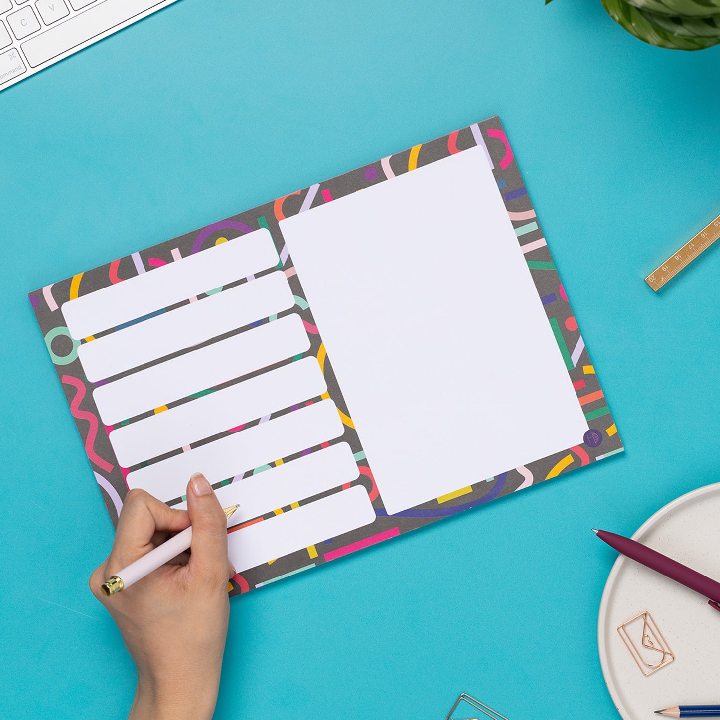 Cutouts A4 Meal/Shopping List Weekly planner pad is on a teal desk, with a hand to its bottom left holding a pen to write on it. There is a white keyboard and paperclips around it.
