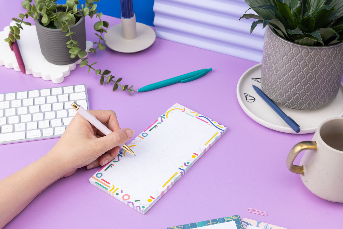 Cutouts List pad is on a lilac desk, with a left hand to its left holding a pen to write on it.  There is a small plant, white keyboard and a mug also on the desk around it.