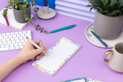 Cutouts List Pad on a lilac desk, with a hand to the bottom left holding a teal pen ready to write on it. 