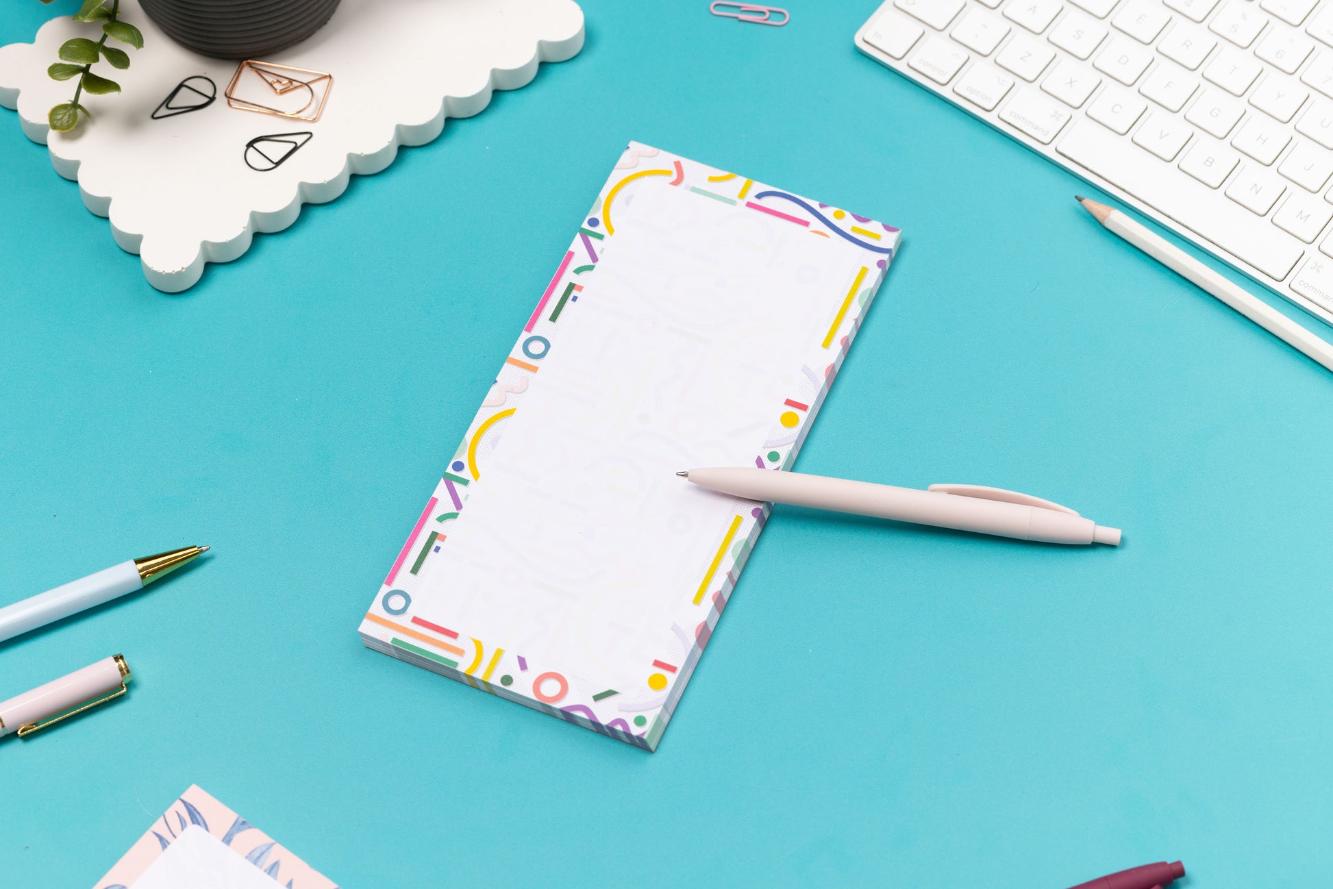 Cutouts List pad is on a teal desk, with a white pen leaning on it, with a white keyboard and paperclips around it.