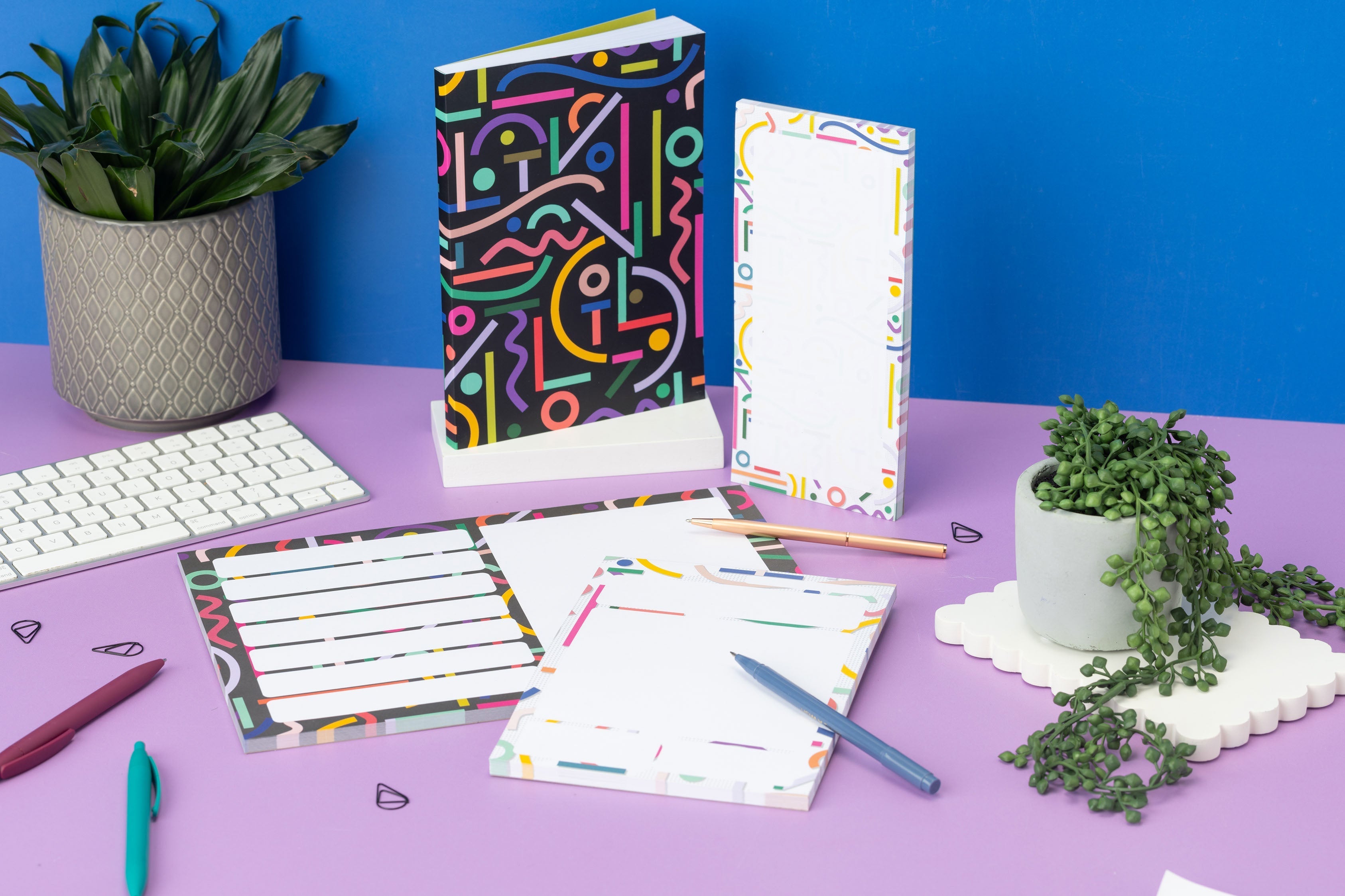 Cutouts Full collection - A4 Planner Pad, A5 Daily Planner Pad, DL List pad and A5 Notebook - are on a lilac desk with small plants, pens and a white keyboard around them. The DL pad is upright, and the A5 Notebook is on a white plinth. There is a blue wall behind.