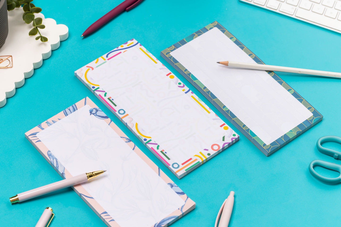 Three list pads - Brighton Birds, Cutouts and Deco Delights - are in a row on a teal desk, with a white pen and pencil leaning on them, with a white keyboard and paperclips around them.