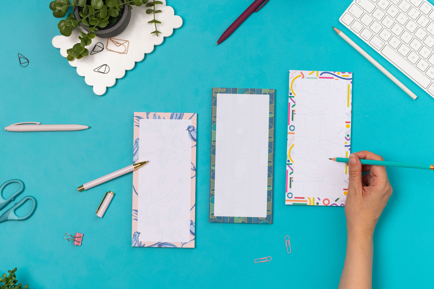 Three list pads - Brighton Birds, Cutouts and Deco Delights - are in a row on a teal desk, with a white pen and pencil leaning on them, with a white keyboard and paperclips around them.