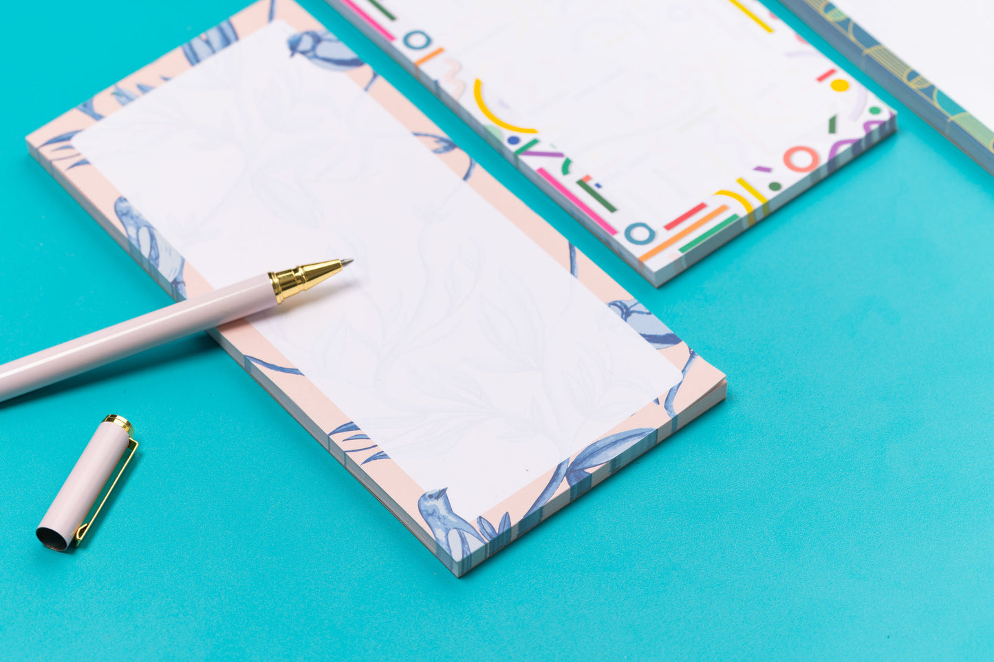 Three list pads - Brighton Birds, Cutouts and Deco Delights - are in a row on a teal desk, with a white pen and pencil leaning on them, with a white keyboard and paperclips around them. The focus of the image is on the Brighton Birds pad, with the other two out of focus to its right.