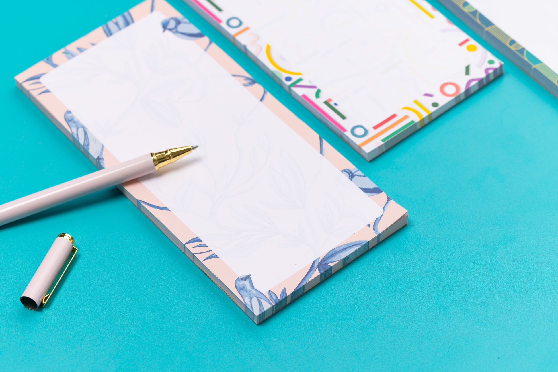 Three list pads - Brighton Birds, Cutouts and Deco Delights - are in a row on a teal desk, with a white pen and pencil leaning on them, with a white keyboard and paperclips around them. The focus of the image is on the Brighton Birds pad, with the other two out of focus to its right.