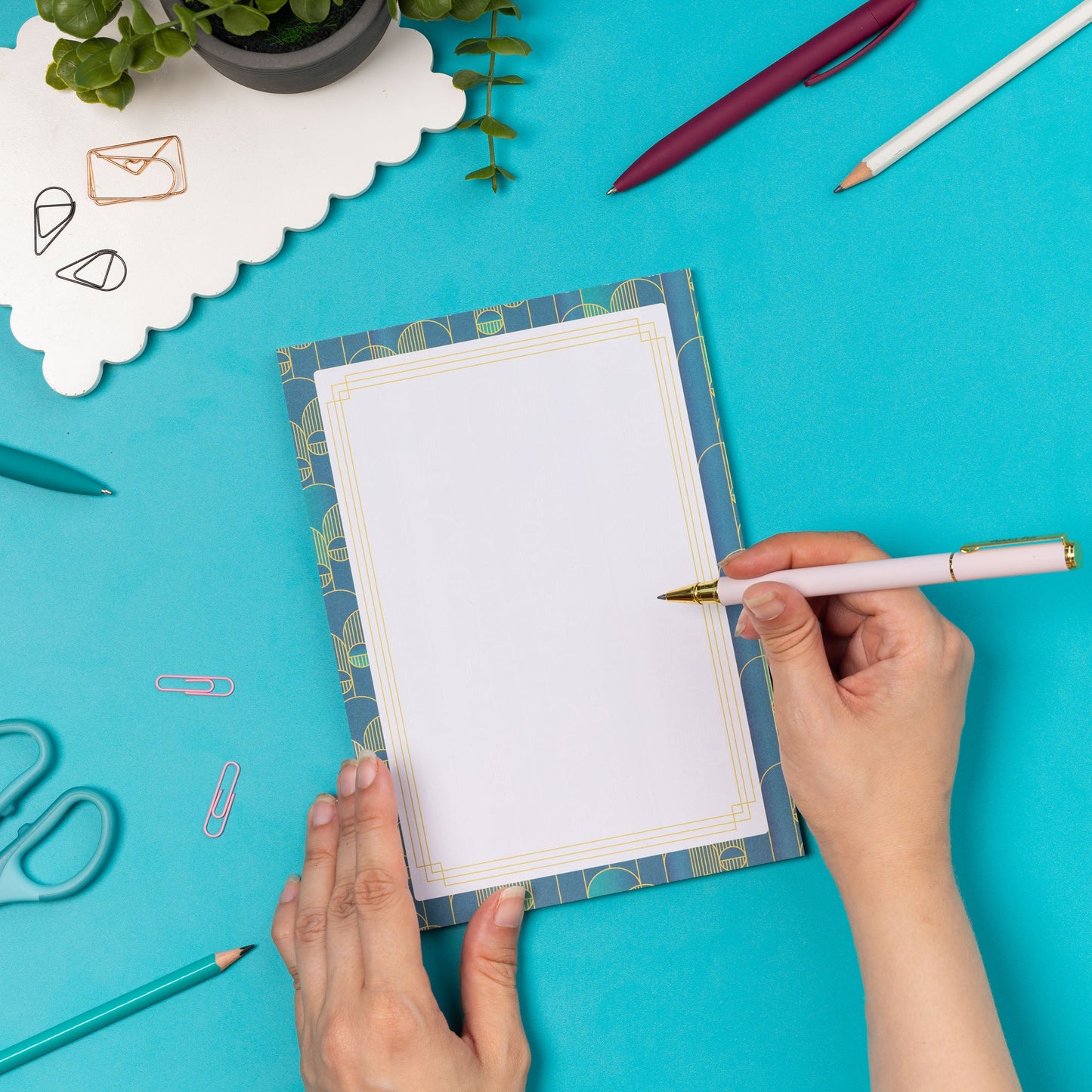 Deco Delights A5 Notepad is on a teal desk with pens and paperclips scattered around it.  The pad is being steadied on the desk to its left by one hand, while on its right a hand holding a white and gold pen is about to write on it.