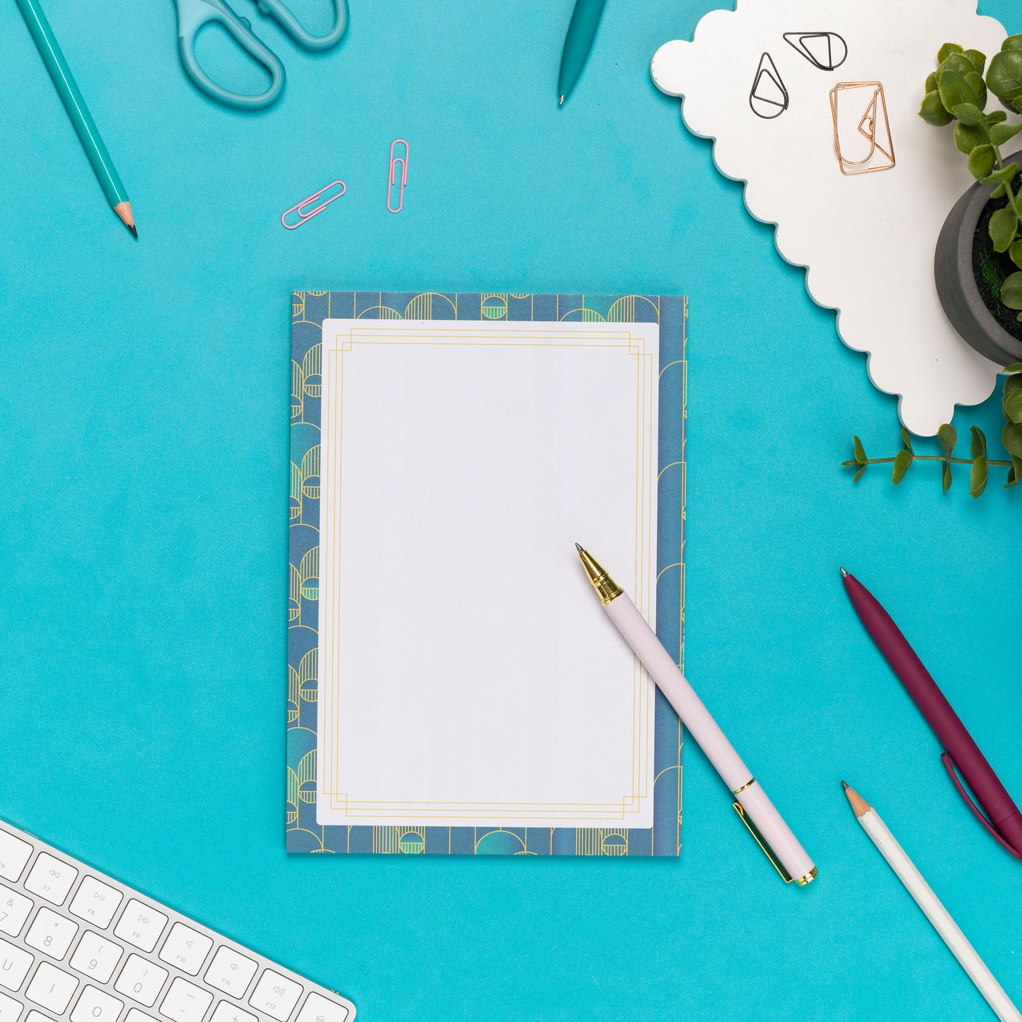Deco Delights A5 Notepad is on a teal desk with a white pend leaning on it and other pens and paperclips scattered around it.  There is also a white keyboard to the bottom left and small plant to the top right of the image.