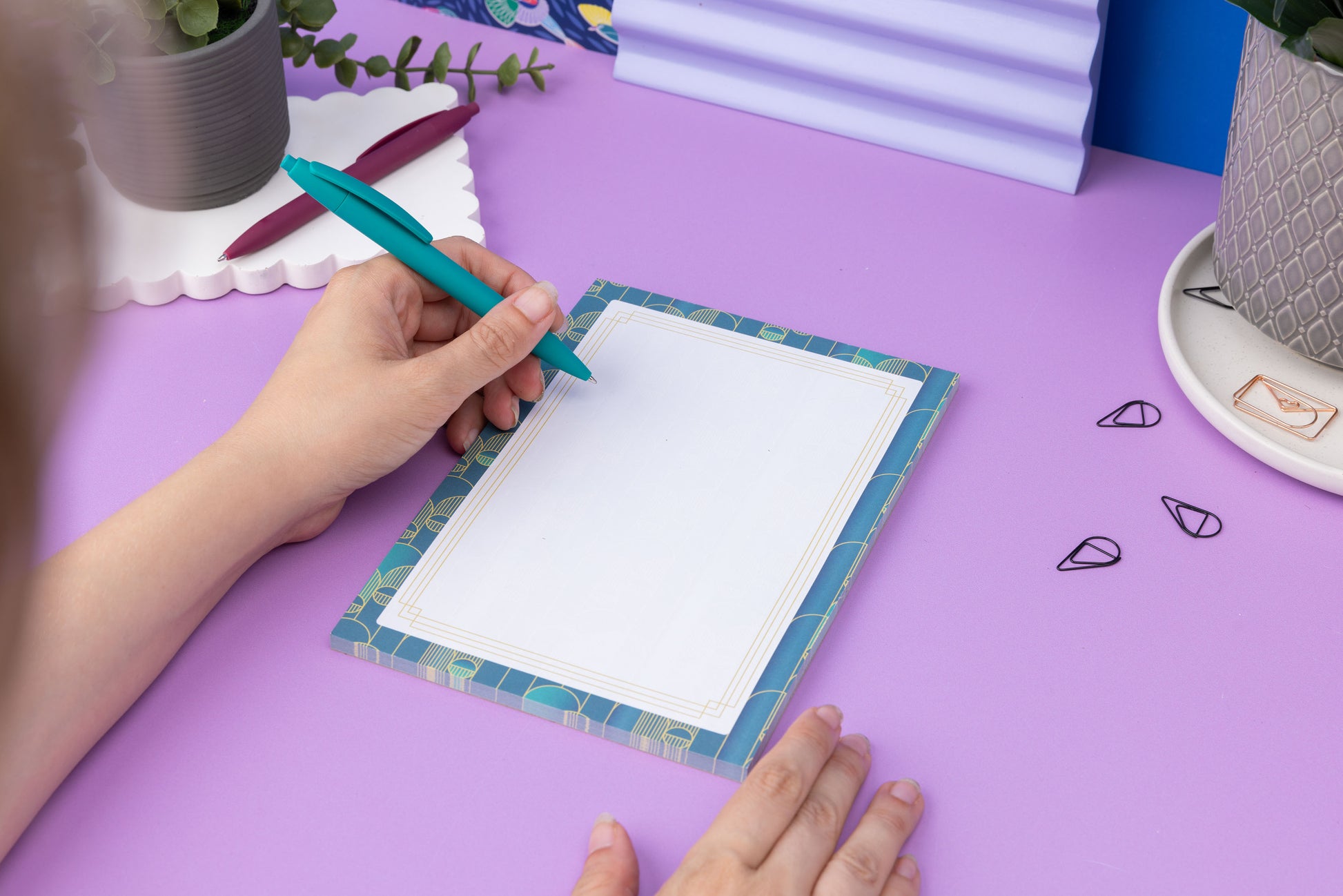 Deco Delights A5 Notepad is on a lilac desk with pens and paperclips scattered around it.  The pad is being steadied on the desk to its right by one hand, while on its left a hand holding a teal pen is about to write on it. You can see the very edge of the writers head in the left foreground out of focus.