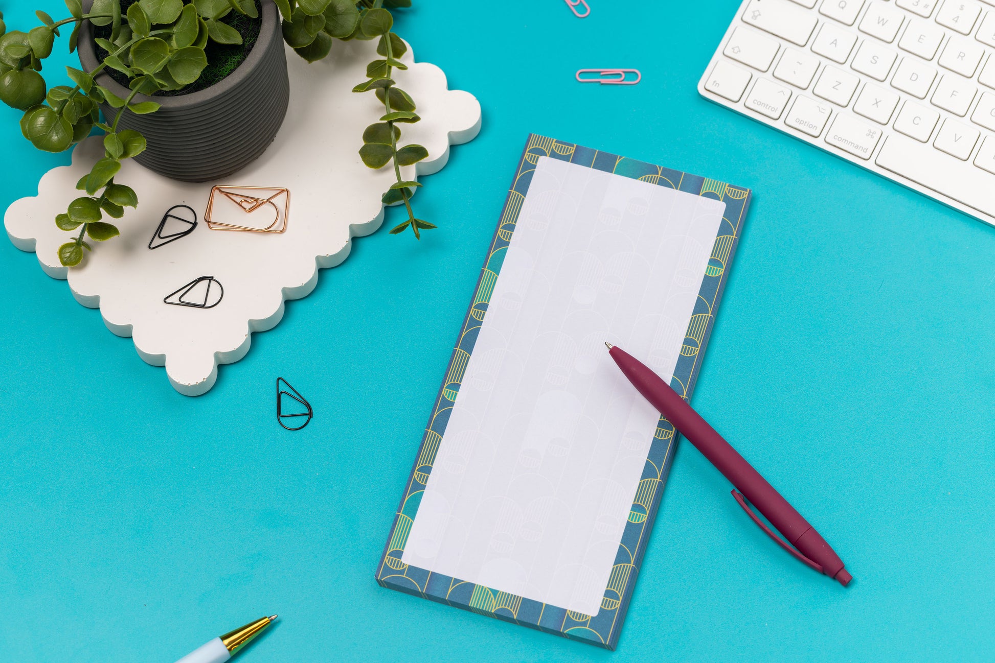 Deco delights list pad - teal gradient background with golden yellow deco motif - is on a teal desk with pen, white keyboard and paperclips surrounding it.