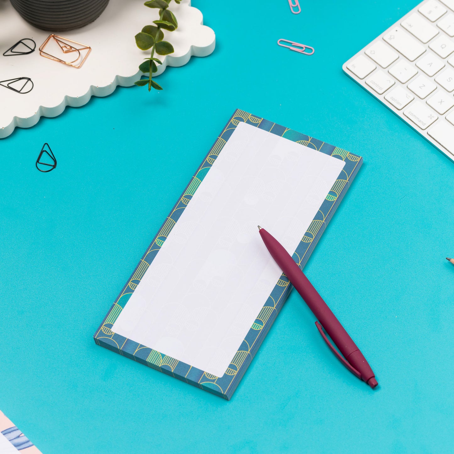Deco delights list pad - teal gradient background with golden yellow deco motif - is on a teal desk with pen, white keyboard and paperclips surrounding it.