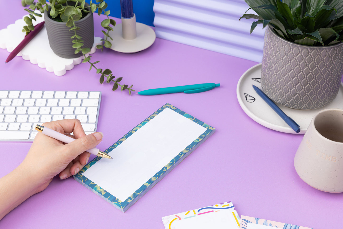 Deco delights list pad - teal gradient background with golden yellow deco motif - is on a lilac desk with pens, pot plants and white keyboard around it. There is a left hand on the left of the image holding a pen to write on the pad.