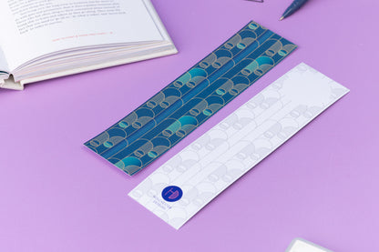 Image of 2 Deco Delights Double sided bookmarks.  One with the full-colour side facing up with the teal gradient and golden yellow deco motif. the other has the monochrome version with the deco motif in a light grey and the Holchester Designs logo at the bottom.  They are on a lilac desk with an open book to the top right corner.