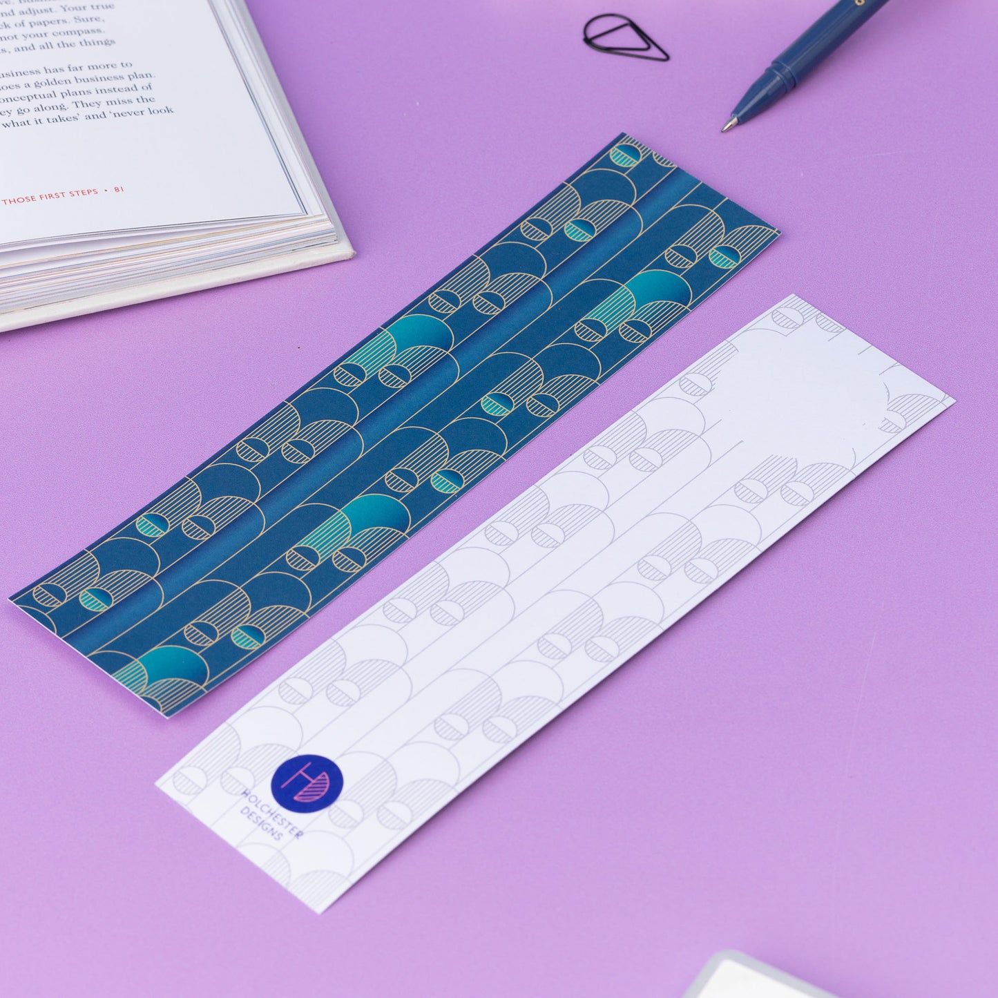 Image of 2 Deco Delights Double sided bookmarks.  One with the full-colour side facing up with the teal gradient and golden yellow deco motif. the other has the monochrome version with the deco motif in a light grey and the Holchester Designs logo at the bottom.  They are on a lilac desk with an open book to the top right corner.