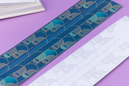 Image of 2 Deco Delights Double sided bookmarks.  One with the full-colour side facing up with the teal gradient and golden yellow deco motif. the other has the monochrome version with the deco motif in a light grey.  They are on a lilac desk with an open book to the top right corner.