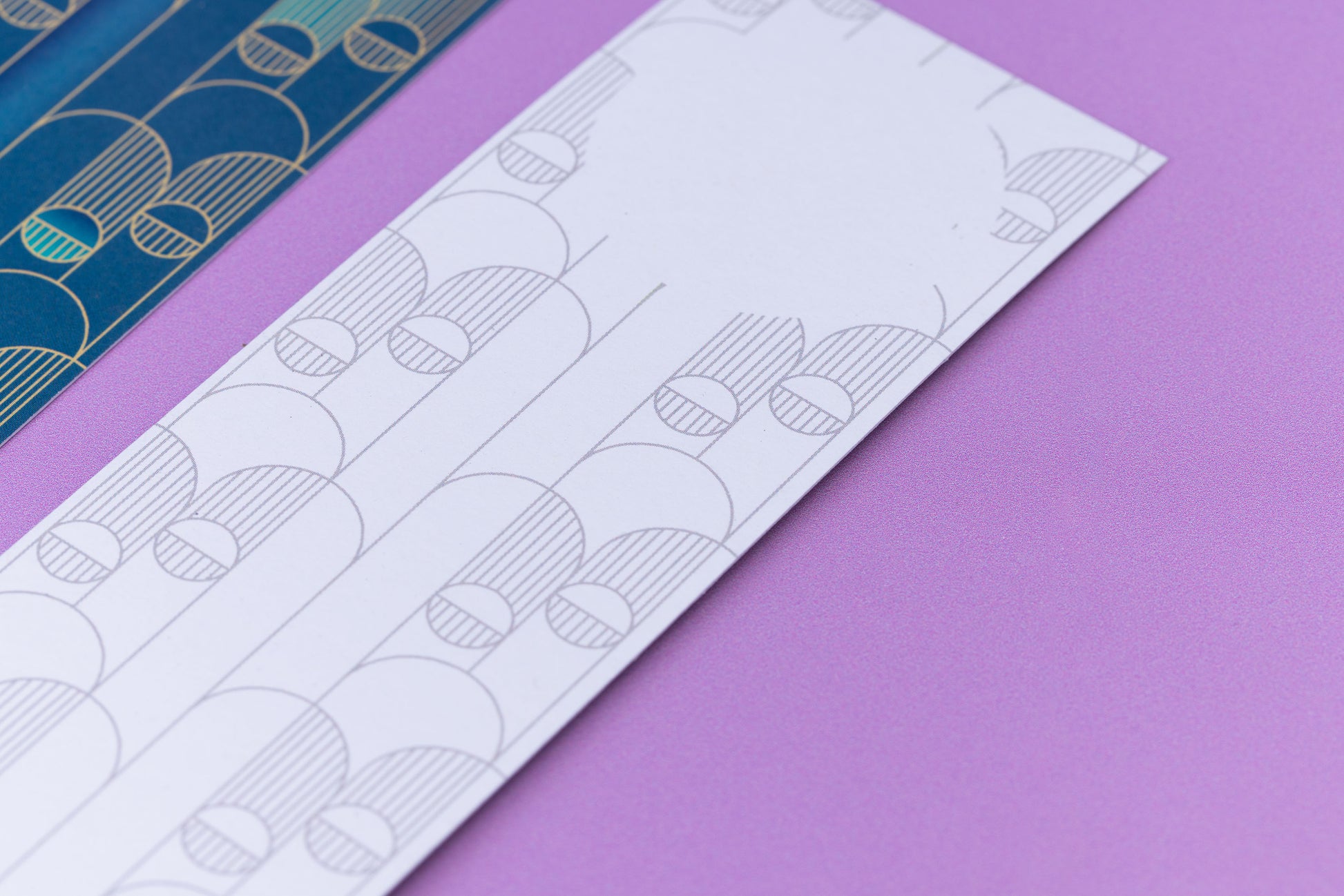 Image of 2 Deco Delights Double sided bookmarks.  The focus is on the top of the monochrome reverse side, showing the blank circle for writing in. Next to it is a full-colour side up, out of focus to the left. They are lying on a lilac desk. 