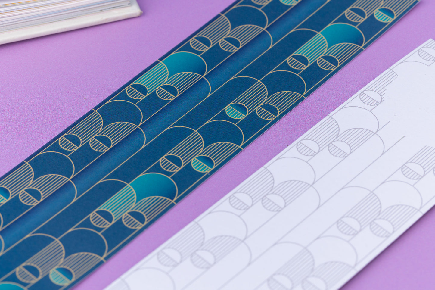 Image of 2 Deco Delights Double sided bookmarks.  One with the full-colour side facing up with the teal gradient and golden yellow deco motif. the other has the monochrome version with the deco motif in a light grey.  They are on a lilac desk with an open book to the top right corner.