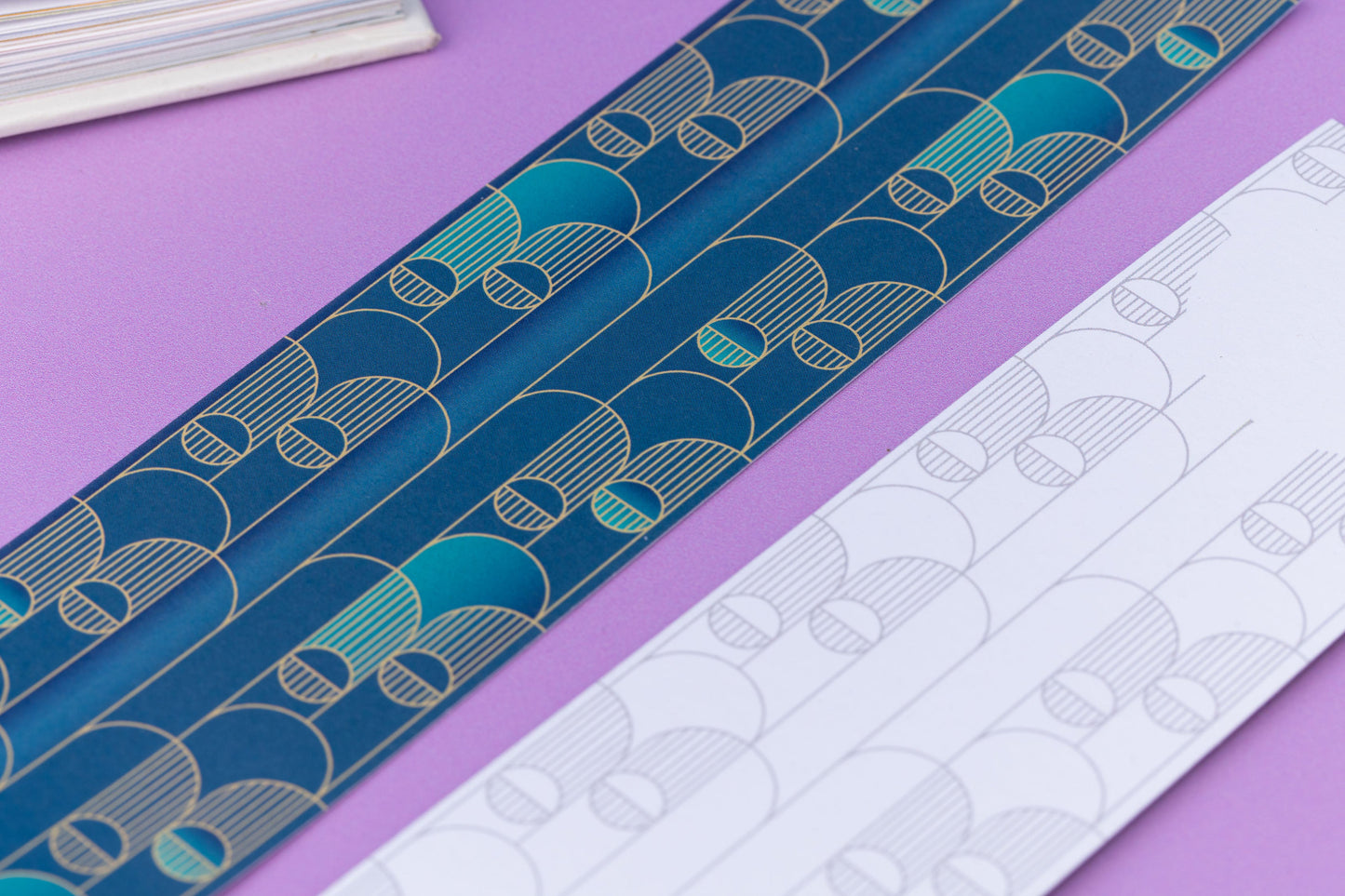Close up of the full-colour patterned side of the Deco Delights bookmark with the teal gradient background and the golden yellow repeat motif.  Next to it is an out of focus corner of the bookmark's other side. They are on a lilac desk with an open book in the top left corner.