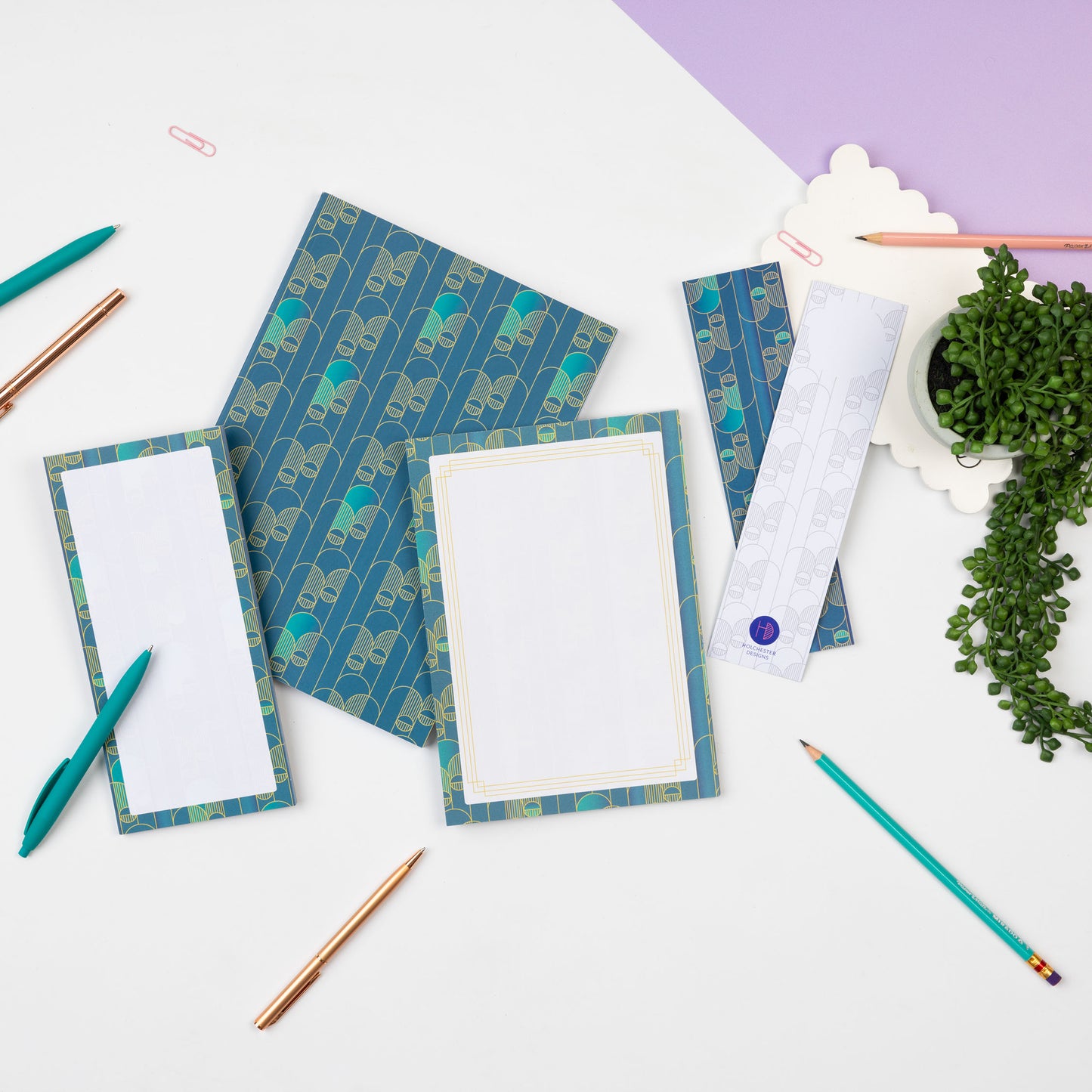 Deco Delights Full collection are laid in a row across a white desk with pens scattered around them and a plant to the right. From left to right there is the List Pad, B5 Dot grid notebook, A5 notepad and two double-sided bookmarks (one of each side facing up).
