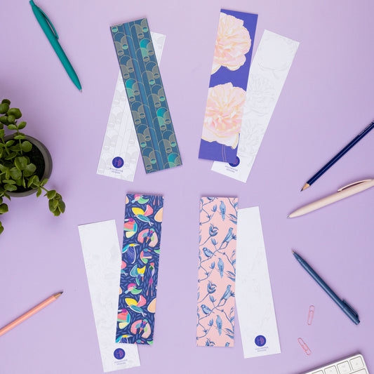 4 pairs of bookmarks are on a lilac table with pens and pencils around them.  Working clockwise there are two of each design - Deco Delights, Spring Forward, Brighton Birds and Paradise - and each have the full-colour version face up on top of the monochrome sides underneath. 