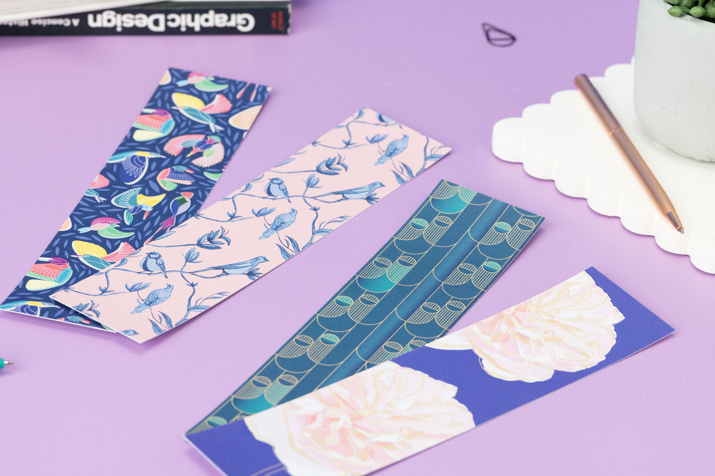 Low photo of the 4 double-sided bookmarks alongside each other, pattern sides facing up, on a lilac desk with books and a scallop edge coaster and plant pot.