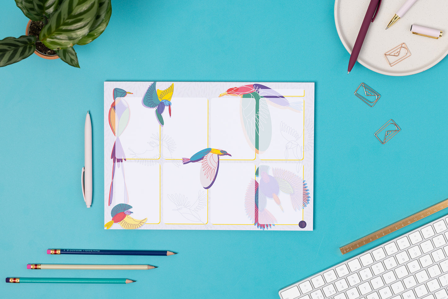 Paradise A4 Planner Pad (8 blocks with bird illustrations around/across it) laid on a teal desk with pens and a white keyboard around it.