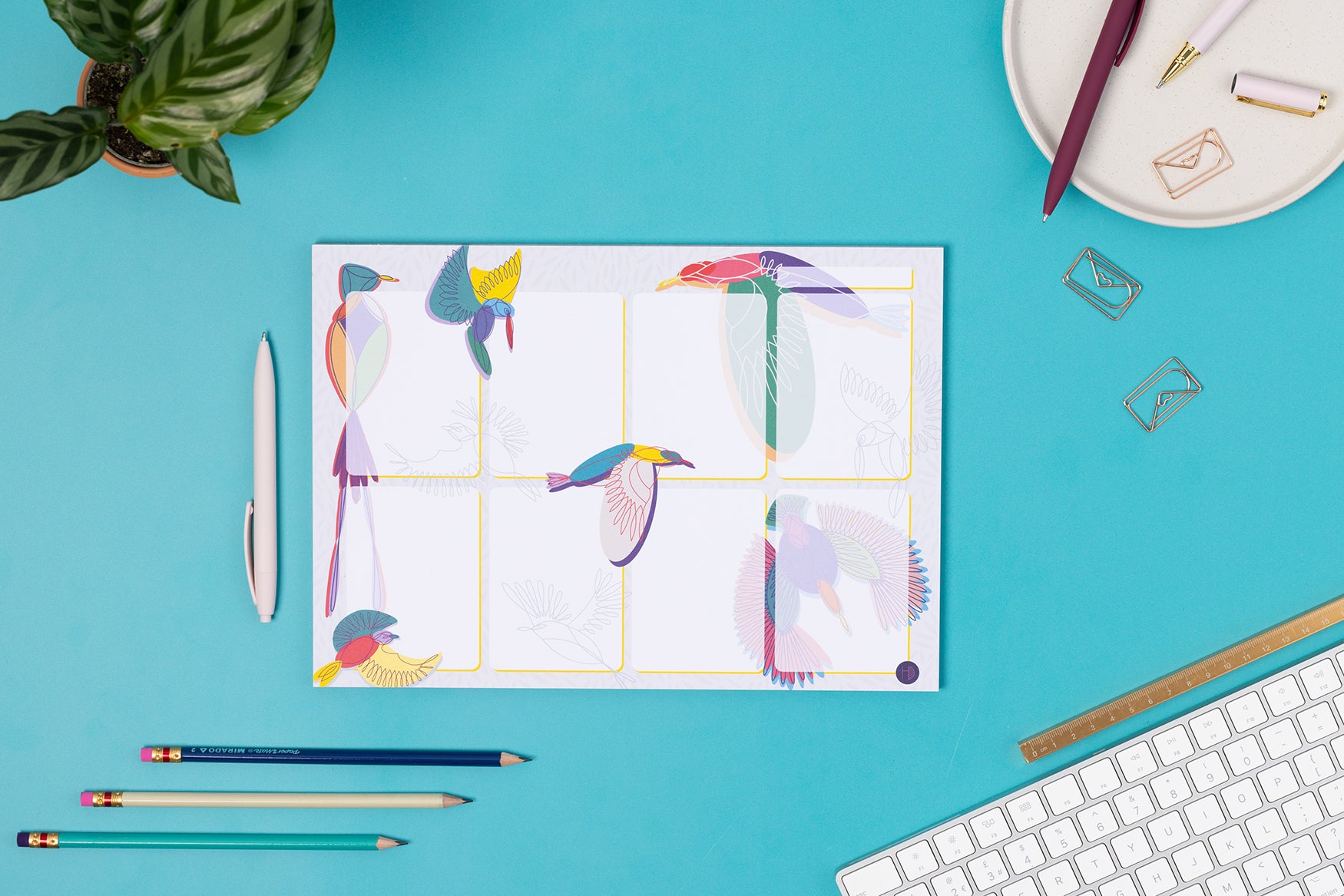 Paradise A4 Planner Pad (8 blocks with bird illustrations around/across it) laid on a teal desk with pens and a white keyboard around it.