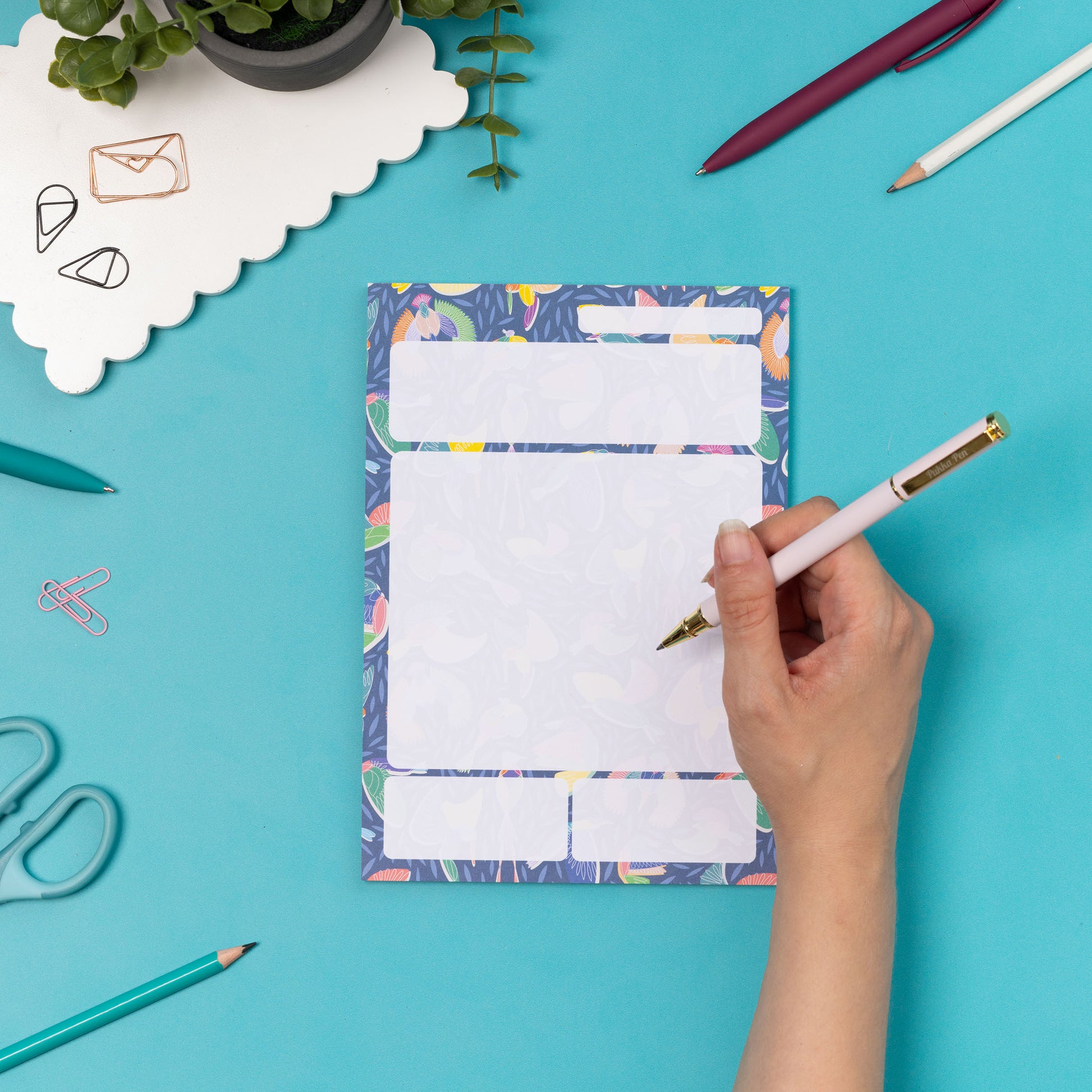 Paradise A5 Daily Planner Pad is central, with a right hand to its right holding a white pen to write on it.  They are leaning on a teal desk, with pens and a small plant around them.