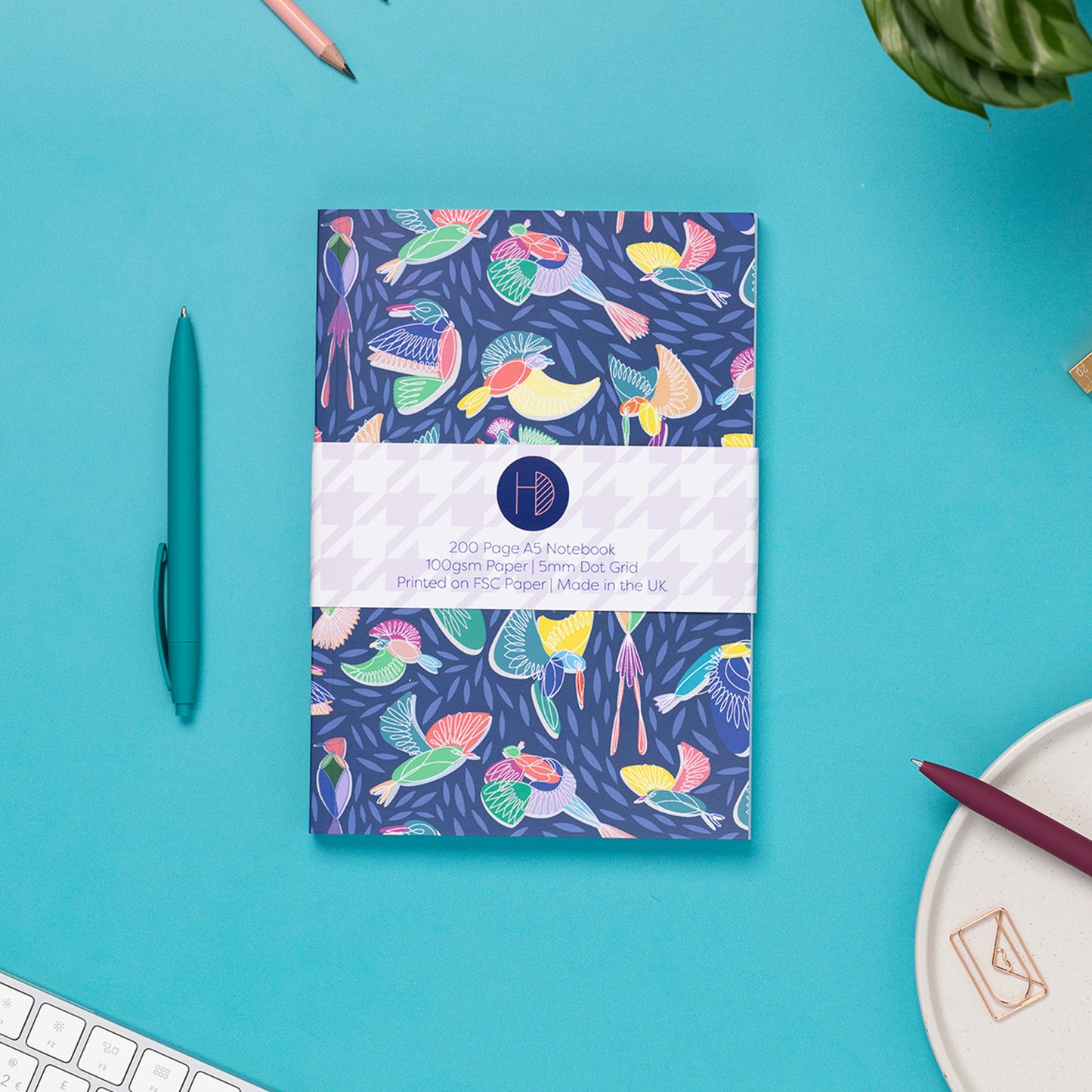 Paradise A5 Dot Grid Notebook, with its belly-band packaging around it, is on a teal desk with some pens, and a keyboard to the bottom left corner.  The book's cover features a navy background with line-drawn bird motifs and offset coloured blocks behind them.