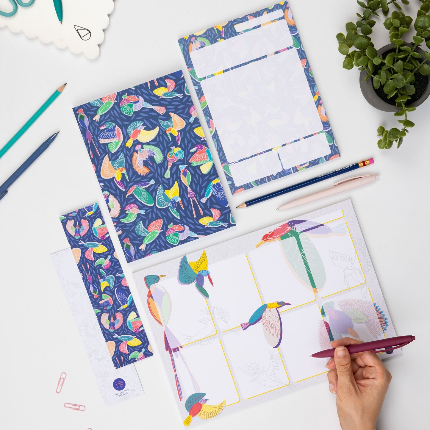 The Full Paradise collection - A4 Planner pad, A5 notebook, A5 Daily Planner Pad and 2 Double-side bookmarks (one side each) - are lying on a white desk surrounded by pens, paperclips and a small plant.  There is a right hand in the bottom right corner holding a pen to write on the planner pad.