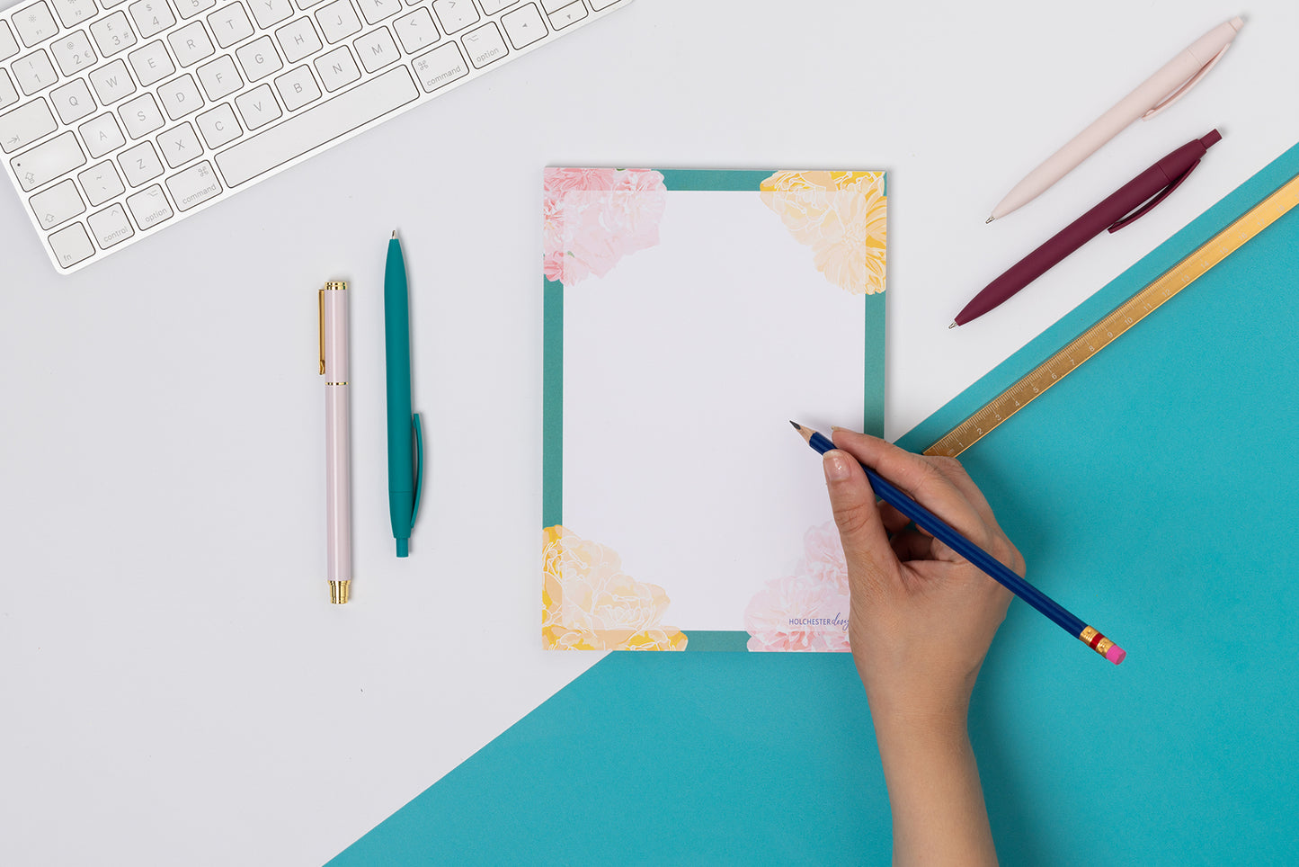 Spring Forward A5 Notepad is flat on a tabletop - half teal and half white cutting across the image diagonally. There is a hand to its right, holding a pencil to write on it, as well as pens and white keyboard around it.
