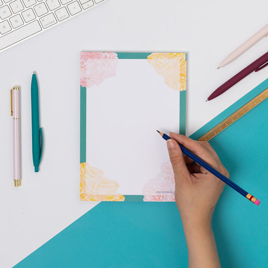 Spring Forward A5 Notepad is flat on a tabletop - half teal and half white cutting across the image diagonally.  There is a hand to its right, holding a pencil to write on it, as well as pens and white keyboard around it.