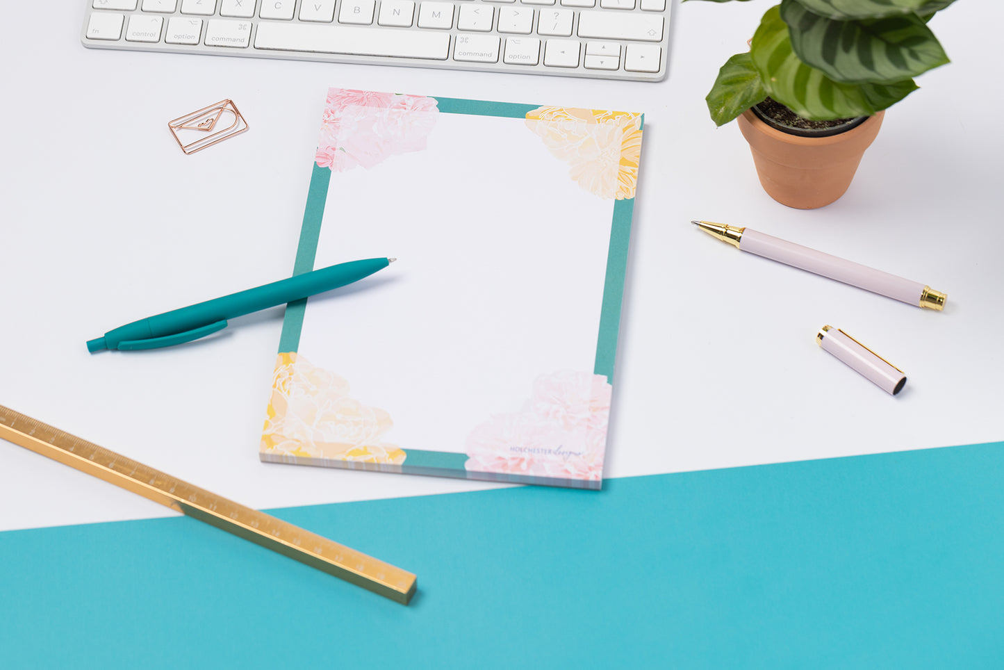 Spring Forward A5 Notepad is flat on a tabletop - half teal and half white cutting across the image diagonally. There pens and white keyboard around it.